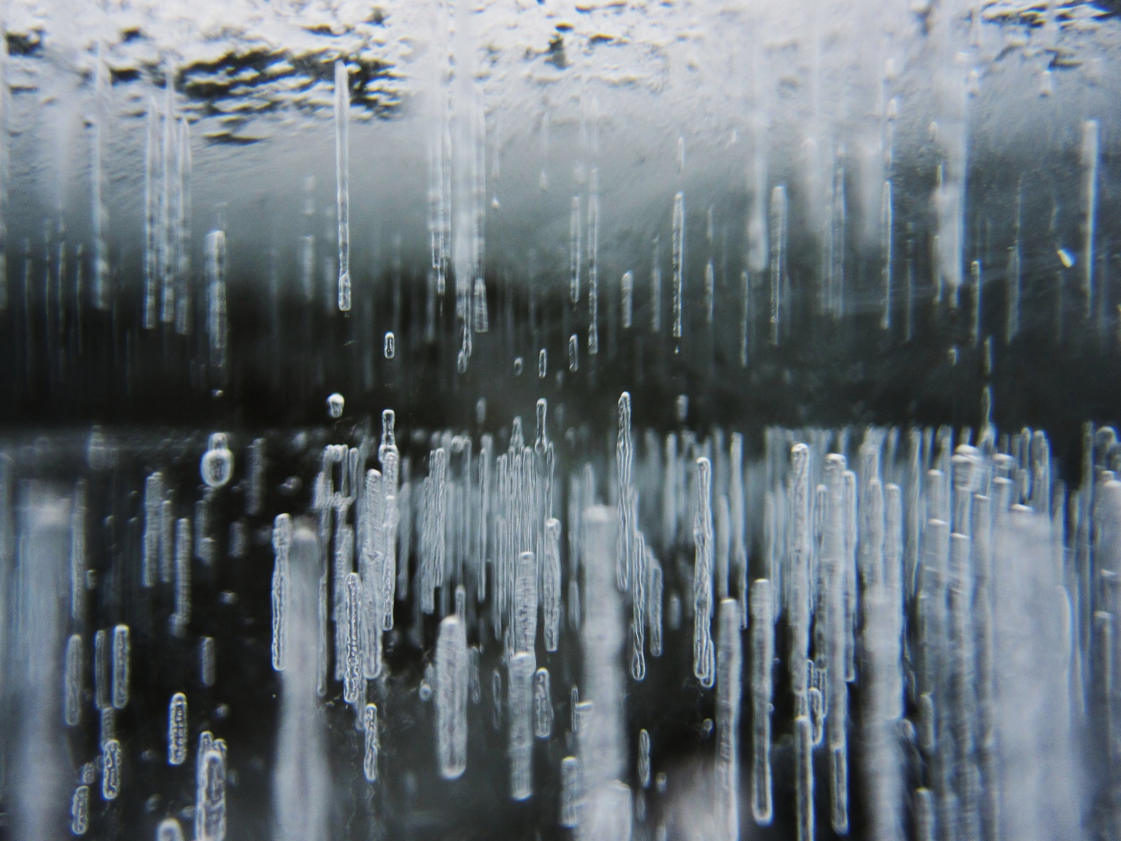 Lake Superior, circa 2010. A macro photograph of air bubbles trapped in ice, forming vertical linear patterns. The bubbles appear as elongated transparent columns of varying lengths suspended within the frozen water, creating a striking natural pattern against a dark background. The cylindrical air pockets are captured in sharp detail, showing how they were frozen in place as the water solidified. Some bubbles are in crisp focus while others blur into the background, adding depth to the image. The translucent nature of both ice and air bubbles creates subtle blue tints and refractions, giving the photograph an ethereal, abstract quality reminiscent of a minimalist architectural drawing.
