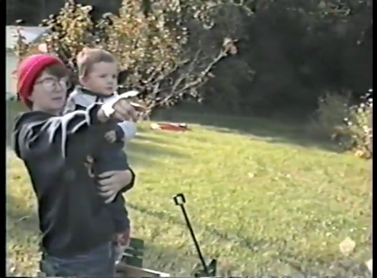 A home video still from what appears to be the 1990s, showing an adult holding a young child, approximately 2-3 years old, in their arms outdoors. The adult is wearing glasses, a bright red knit cap, and a dark sweater or jacket. The child is wearing light-colored clothing. They're standing in what looks like a backyard with grass and autumn trees or bushes with yellowing leaves in the background. The image has the slightly fuzzy quality characteristic of vintage home video footage.
