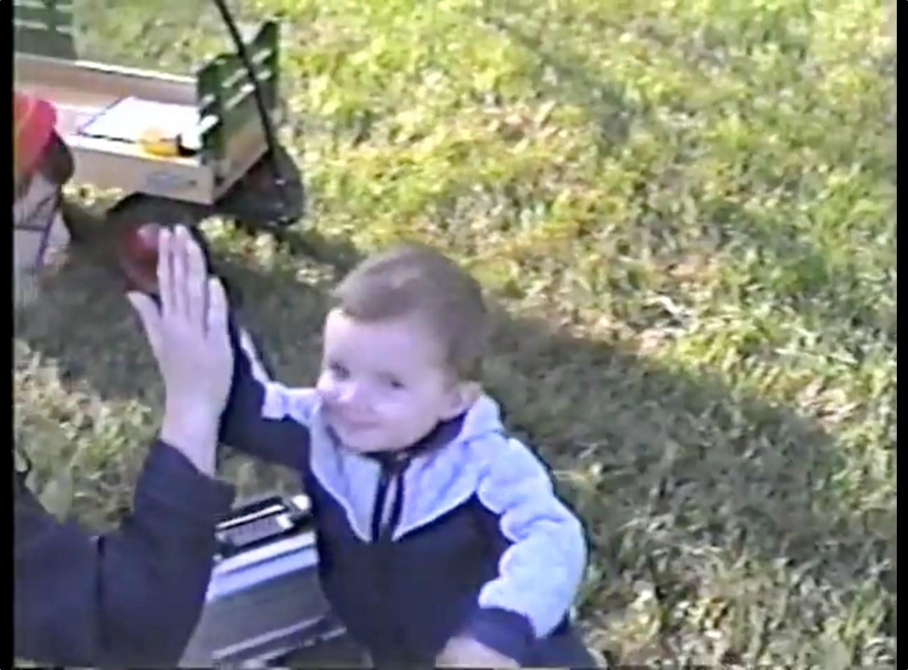 A heartwarming moment from the same home video where a toddler is smiling and giving a high-five the same adult off-screen. The child is wearing a two-toned outfit with a light-colored (possibly white or light blue) top portion and darker bottom. The scene is still set in the yard, with sunlit grass visible in the background. The video has that characteristic VHS quality, giving it a nostalgic 90s feel. There appears to be some kind of box or container visible at the edge of the frame.